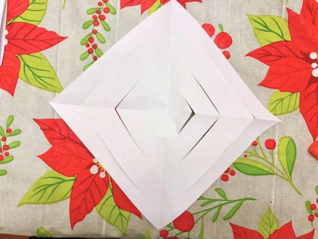 square piece of white paper sitting with corners at top and bottom and left and right (positioned like a diamond) with straight cuts throughout, sitting atop a poinsettia design tablecloth