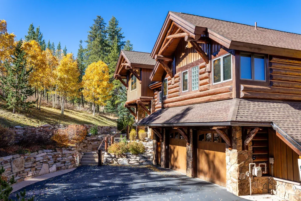 exterior of a two story home nestled among trees changing colors.  The exterior is logs as the facade on the 2nd story and 2 wood garage doors on the first floor.