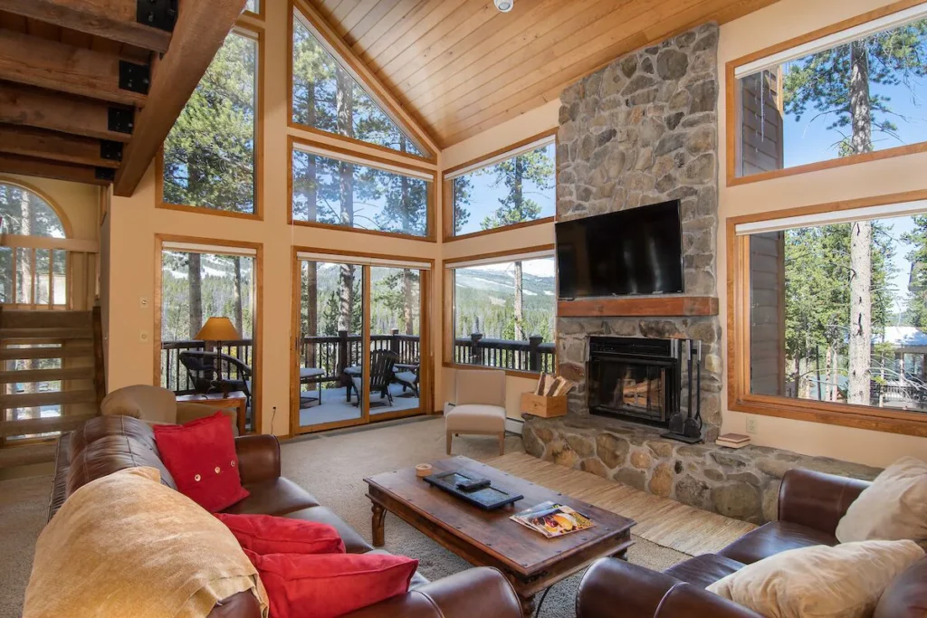 a large living room with a floor-to-ceiling stone fireplace, floor-to-ceiling window, couches with red decorative pillows, and pine trees and mountain views beyond the windows in this Breckenridge rental home