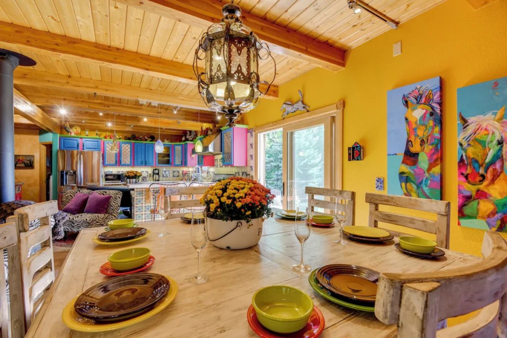 inside a rental home in Steamboat Springs. Vibrant yellow/orange walls, a large wooden dining table with wooden chairs and colorful place settings. The kitchen in the back has cabinets painted in bright blue and pink hues.