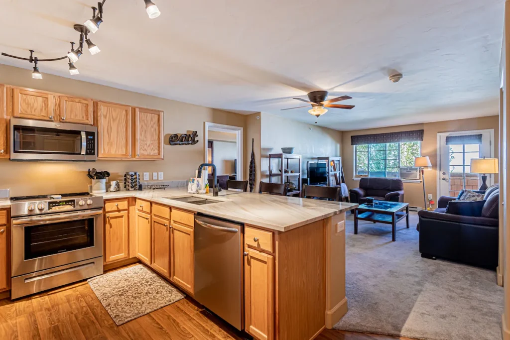 looking through a small kitchen area with a peninsula counter, then into a living area. Lights are on, cabinets are light oak, and living area is carpeted