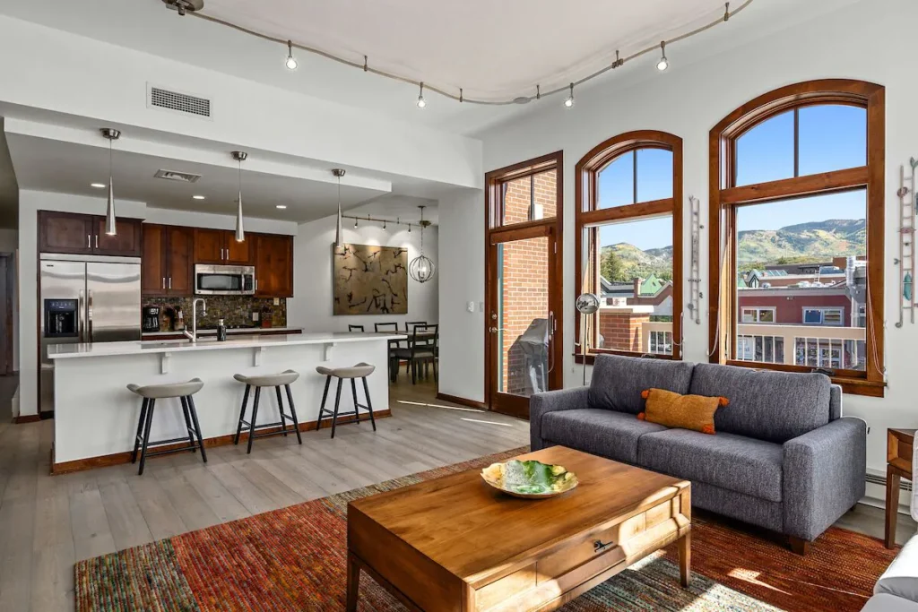 a condo rental in Steamboat Springs right in town. A kitchen with a long island counter and 3 white stools, large wood-framed windows to the right on the wall look into town buildings, a living area in the foreground with a gray couch to the right and wooden coffee table in the middle