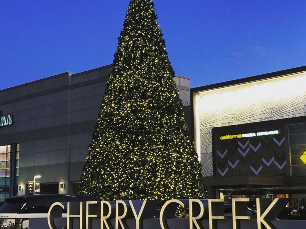 tall christmas tree in front of a mall building.  Capitol Letters that say "CHERRY CREEK" lit up in front of the tree.  Sky is getting dark.