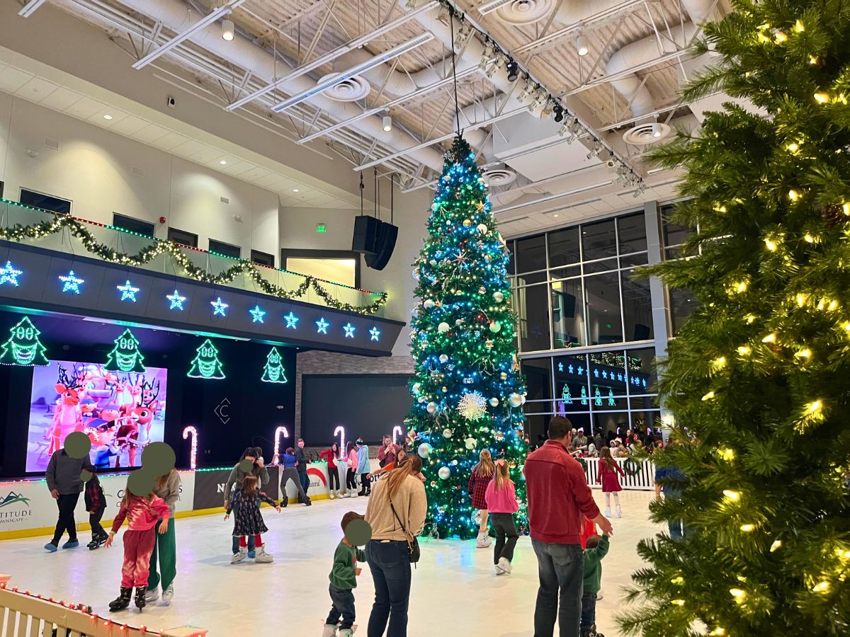 an indoor ice skating rink with a tall tree in the middle, rudolph video playing on a large screen to the left, and plenty of kids and adults on the ice