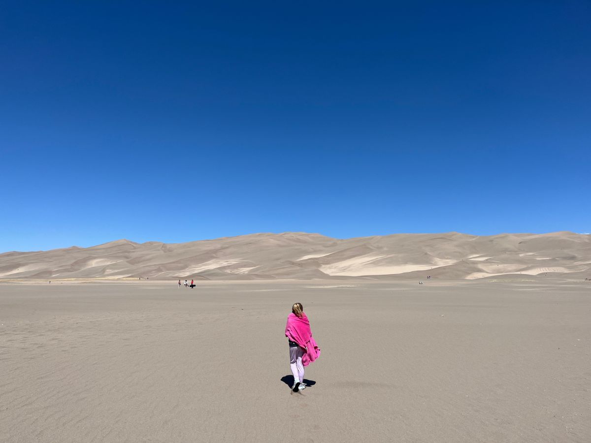 Great Sand Dunes National Park. A young girl wrapped in a dark pink blanket walks on the flat sand toward the taller dunes on a clear blue sky day