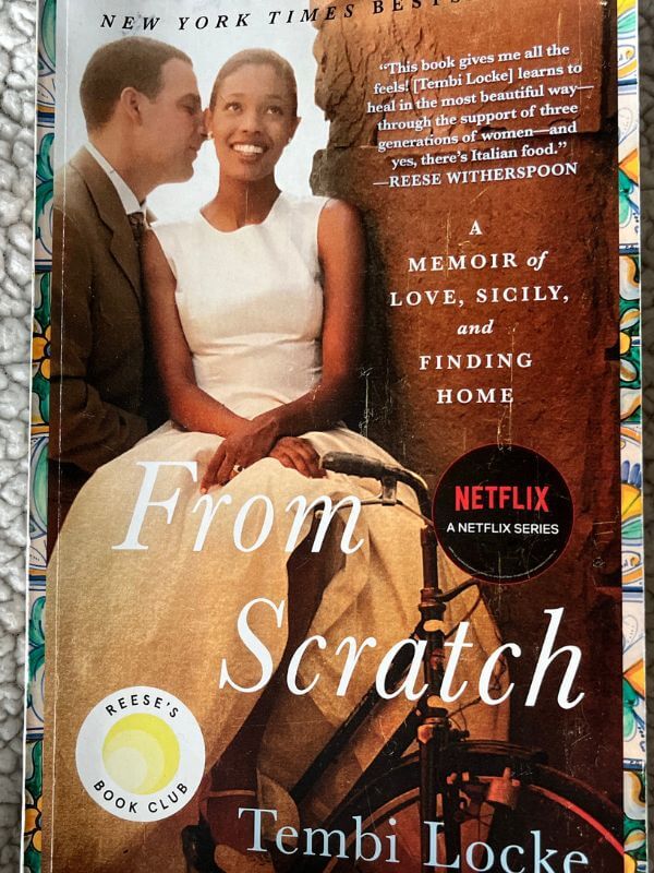 cover of book From Scratch. A couple sitting in a window ledge, girl in white top and flowing yellow/beige skirt looking forward, boy turned to see his profile, about to kiss her on the cheek or whisper something in her ear