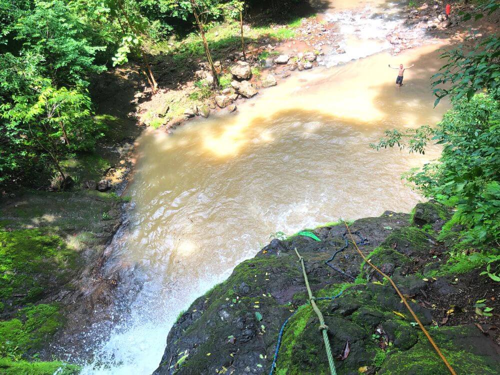 Looking down from the top of Salto de Gamalotillo waterfall. A man stands at the bottom in the brown water with his arms spread out, a few thin ropes hang down the side of the cliff