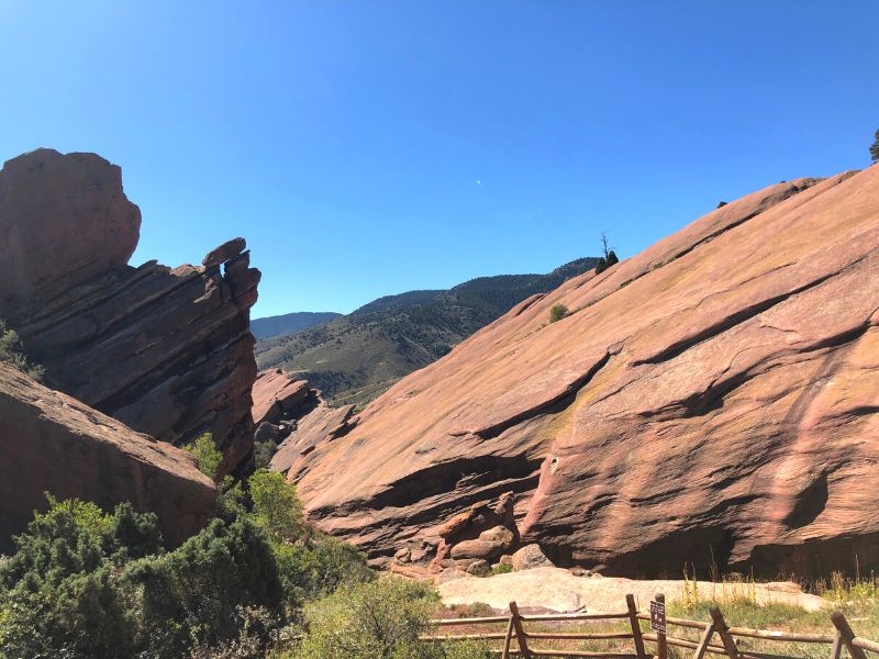 Red Rocks scenic spot, smooth and tall red rock wall, fence in foreground, greenery in bottom left, blue skies
