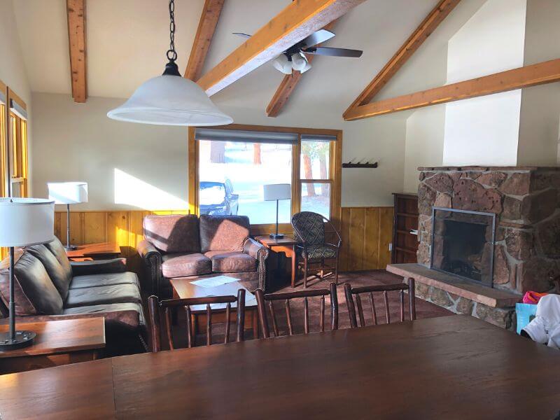 indoor cabin area, overlooking large dining table into the living area with stone fireplace to the right and tall ceilings with wooden beams