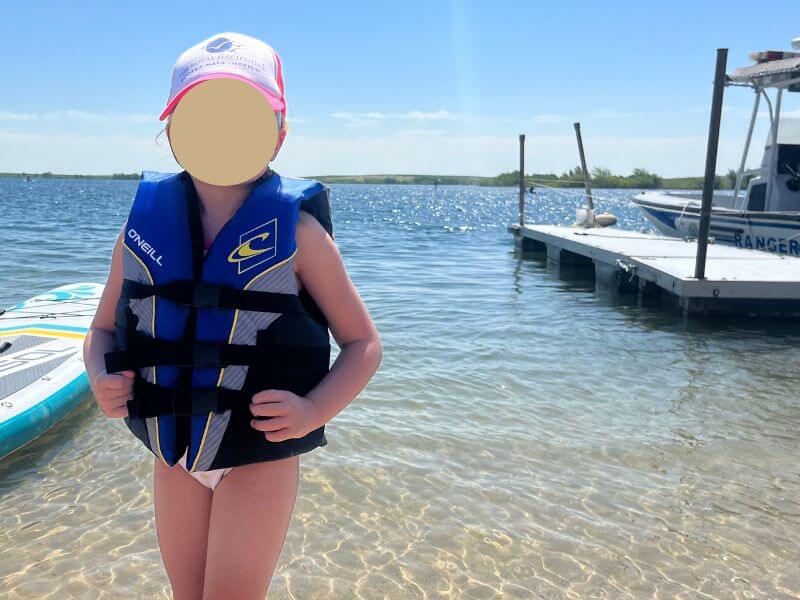 little girl standing at edge of lake, murtisol paddle board at the side, she is wearing a blue life jacket
