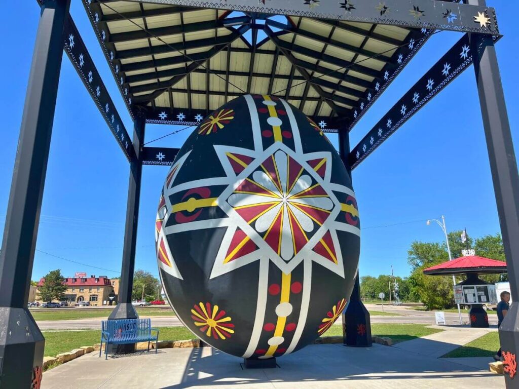 A giant Czech egg under a covered area. Egg is mainly black with white stripes and a white flower in the middle, symmetrical design with red and yellow accents