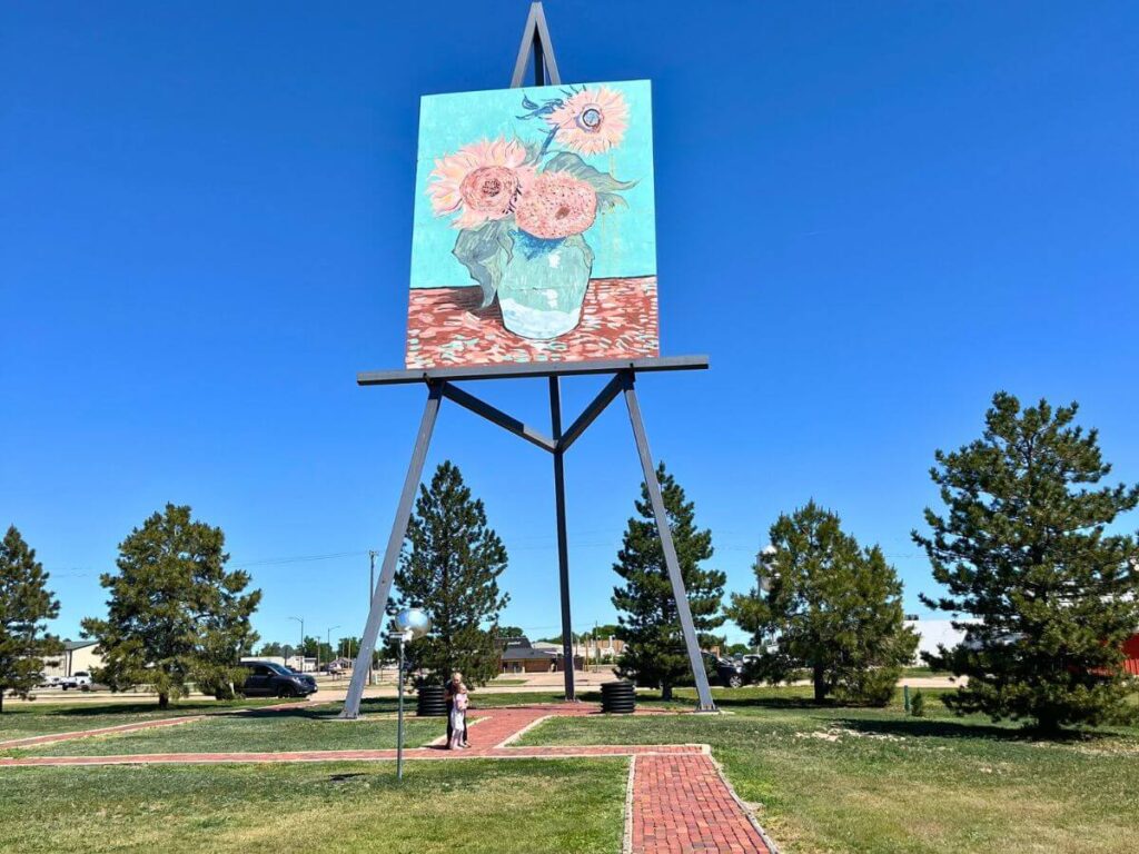 a giant, very tall easel with a huge painting of VanGogh's 3 flowers all set in a park area of green grass and a few evergreens in the background