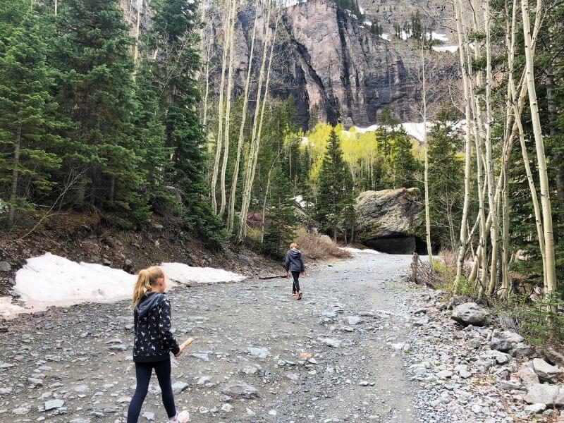 two kids on a rocky dirt trail, snow on the side and large boulder ahead