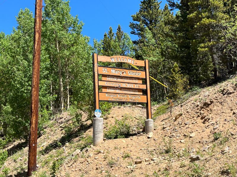 wooden sign on side of road stuck in dirt/mountain welcoming you to St. Mary's Glacier, evergreens behind