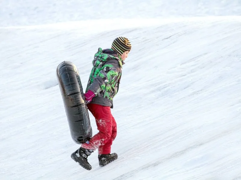 boy in red pants carrying a black snow tube up a snowy hill