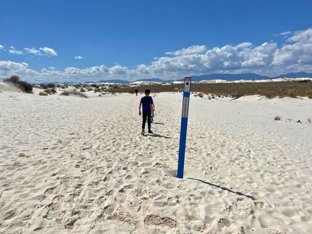 boy walking away in white sand with a post marking the trail in the foreground, dry plants in the distance, blue skies with a few clouds