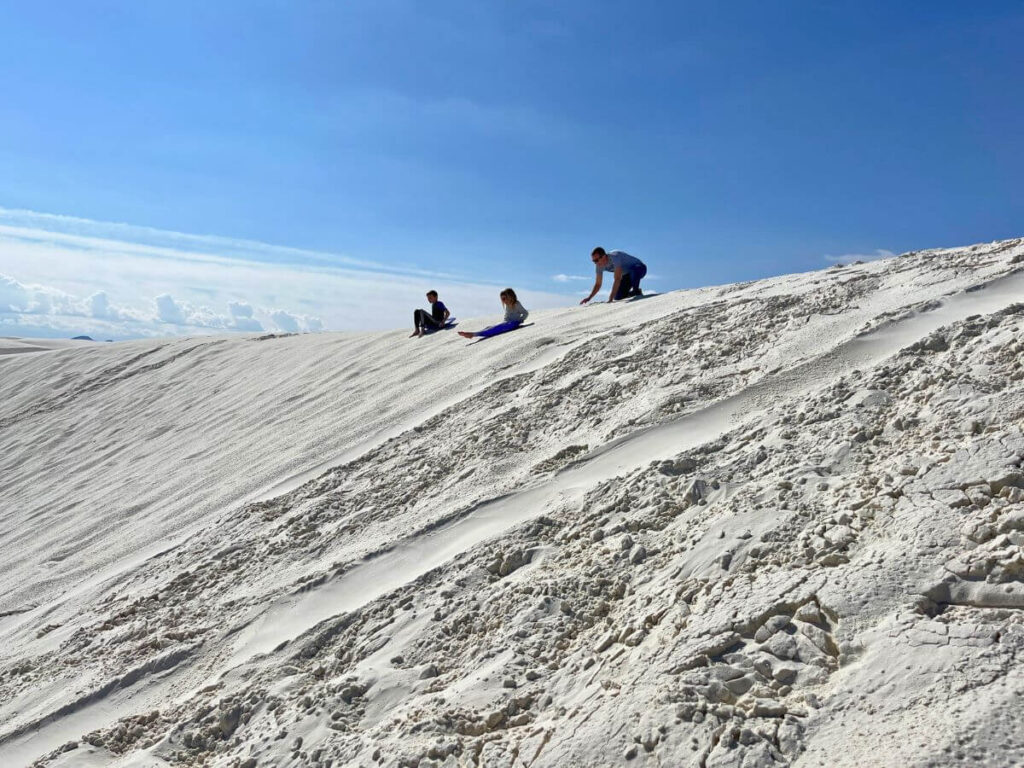 kids at a distance sitting on sleds at the top of a white sand hill, blue skies in the back