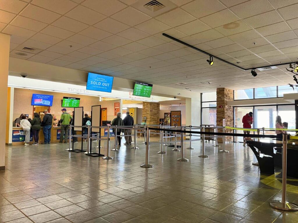 indoors of the Carlsbad Caverns visitor center, a line queue that isn't filled much, white tile floor, tv's hanging above with check in staff behind a counter at the front of the line