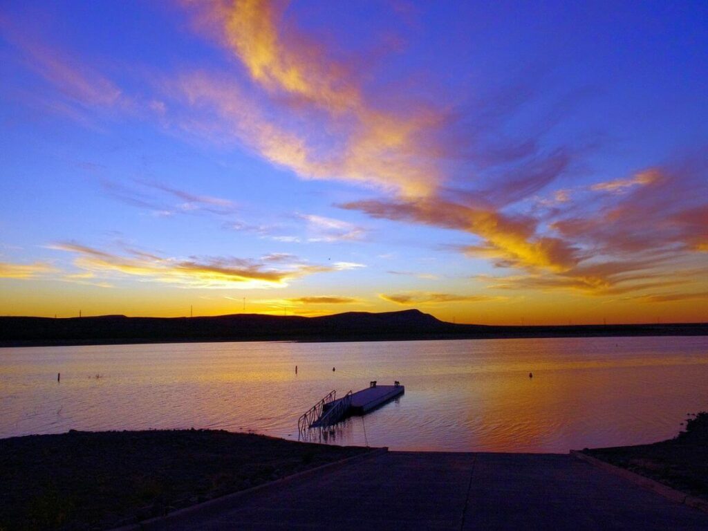 a lake at sunset, a small dock in the water, tinted blue sky with a streak of a pink cloud coming near the viewer