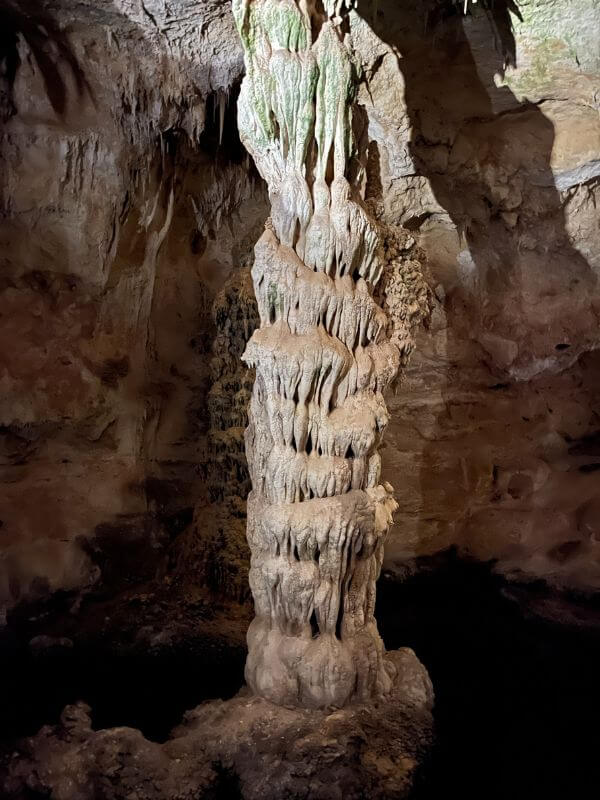 inside Carlsbad Caverns, a dark room with light shining on a tall, skinny formation that is a light beige color and appears to be "dripping" with hundred of dripping rocks