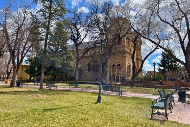 green grass area with a bench to the left, paved walkway to a large cathedral in the background, with a few bare trees in front