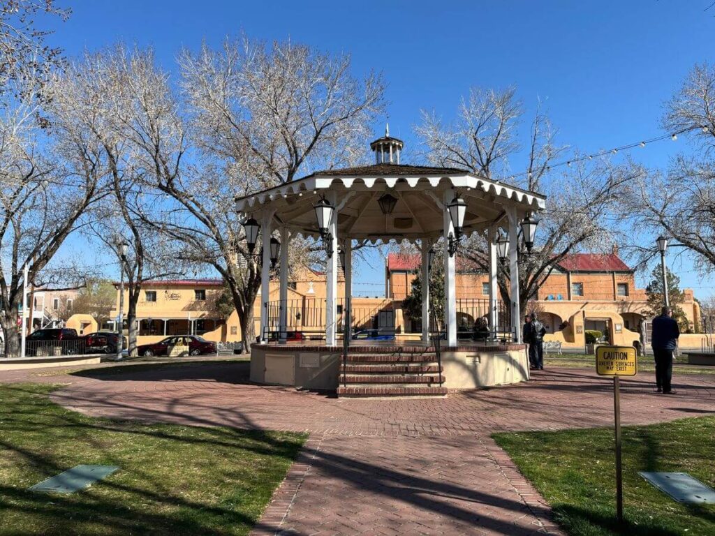 Old Town Albuquerque has a central plaza with a gazebo in the center, paver walkway around the gazebo, grass area to the sides of the walkways.  Bare trees sporadic around the plaza and tan colored buildings in the far back