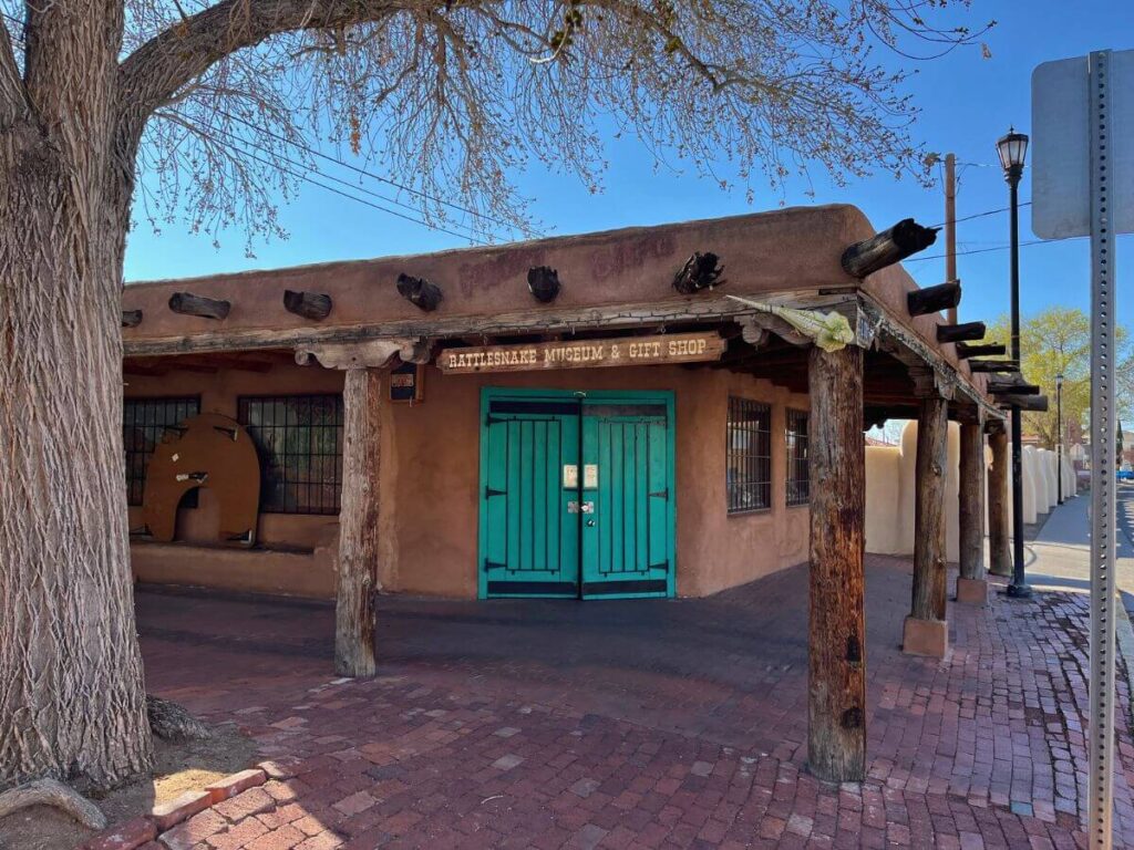 exterior of rattlesnake museum in Albuquerque New Mexico.  A one-story, flat roof adobe building with turquoise double doors. Natural-looking wooden posts to hold a covered area around the exterior walk