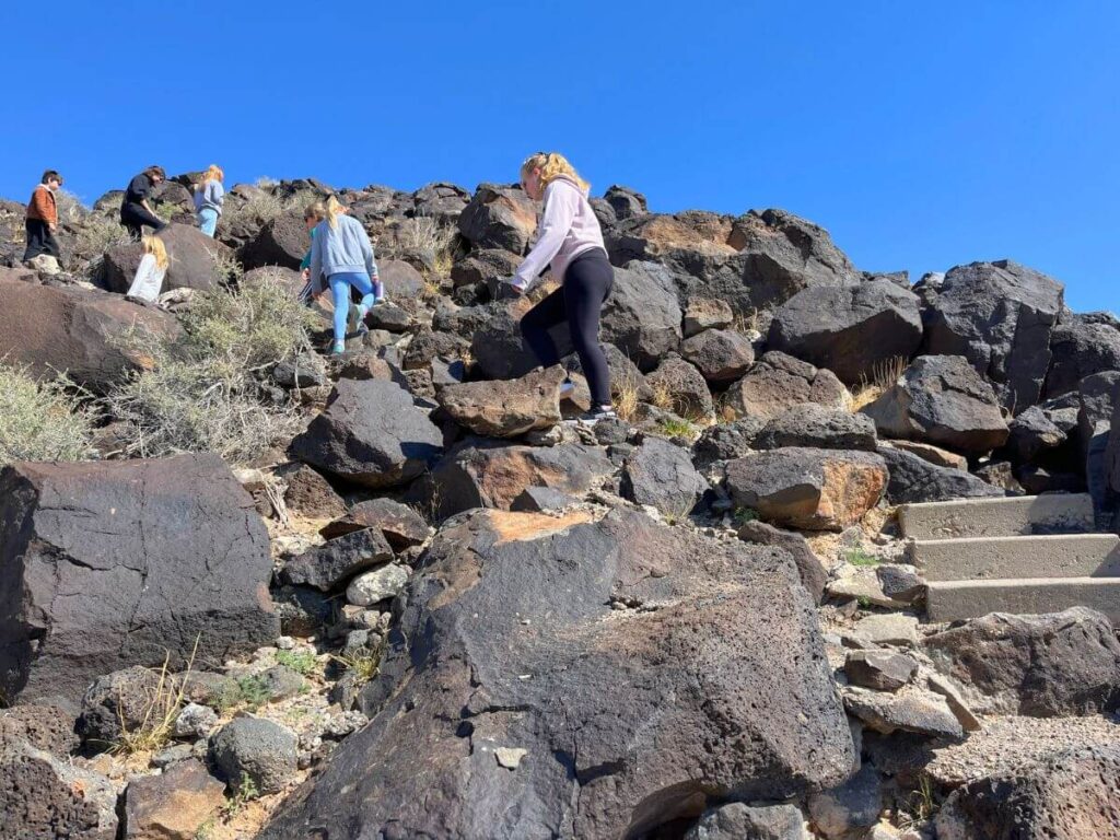 kids climbing up steep rock area, large dark boulders, blue sky above