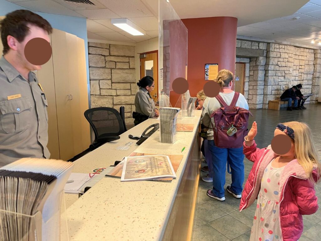 Carlsbad Caverns with kids includes completeing the junior ranger program: a little girl in a pink jacket and white dress with colorful polka dots with her hand up repeating an oath to a park ranger behind a desk for the junior ranger program at Carlsbad Caverns