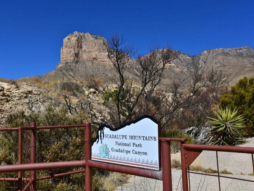 large mountain in back with "Guadalupe Mountains National Park - Guadalupe Canyon" sign in the foreground and a brownish red rail protecting a drop off