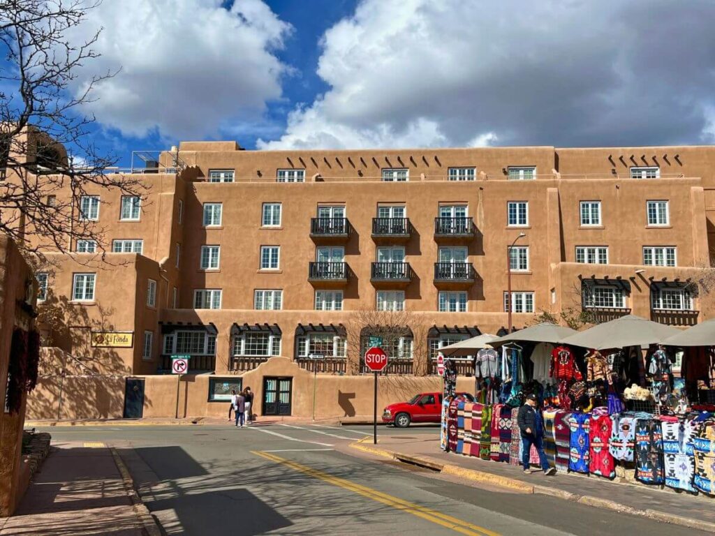 multi-story hotel from the outside, adobe style with tan colored exterior.  Foreground is a road with artisans selling goods on the right