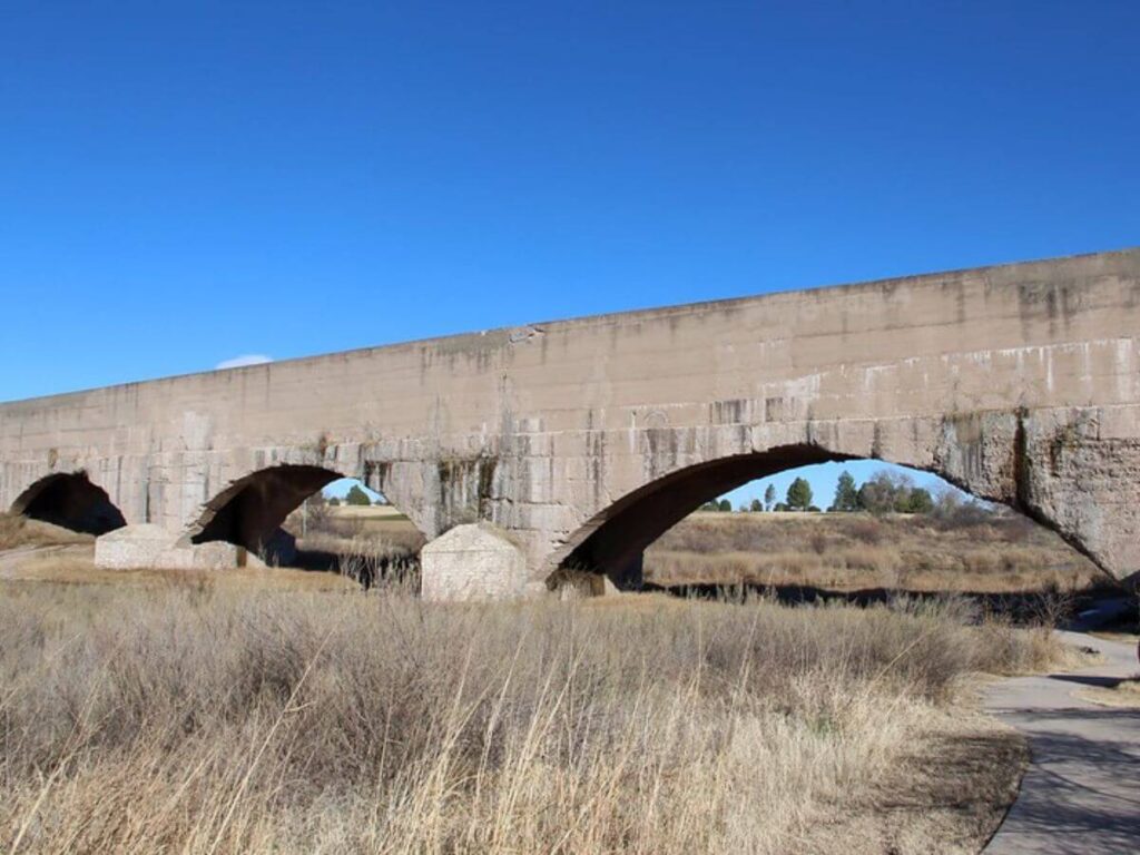 large concrete flume over dry grasses.  Flume has 3 arches in picture and flat on top.