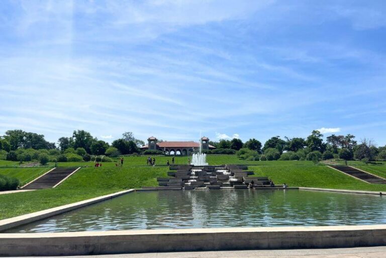 a manicured shallow water area with a grand fountain in the distance at the start of the water, bright green grasses all around, the World Fair Pavillion in the far distance at the top of the hill behind the fountain
