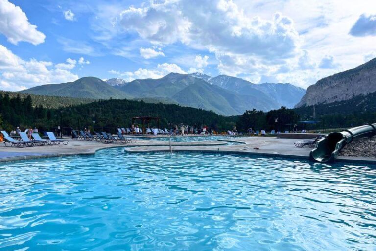 an aqua colored pool with a green tube slide dropping into it on the right. A narrow winding walkway in the water to another section of pool beyond the first one. Pool loungers on the left side and mountain silhouettes in the backdrop