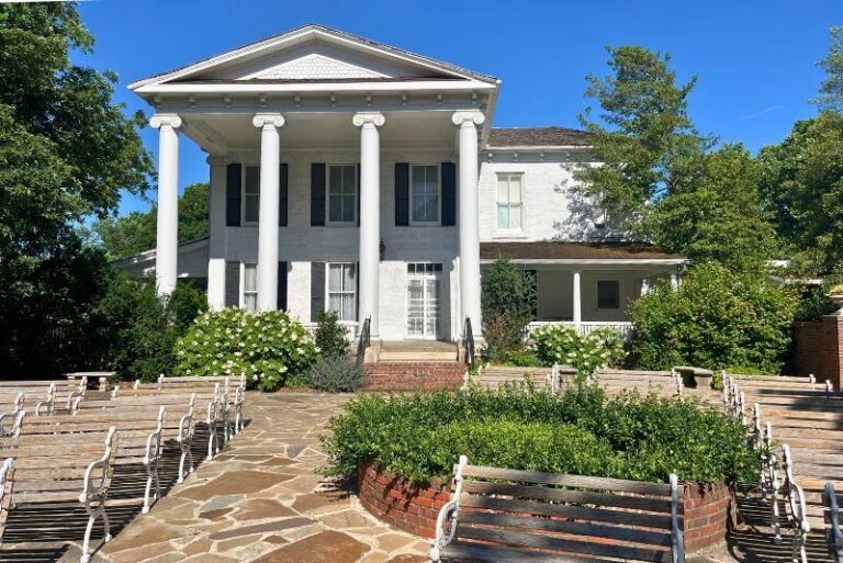 a historic white mansion with 4 2-story columns on front porch, windows with black shutters. Wooden benches lined up as if for an even in foreground, with plenty of trees in background around the house