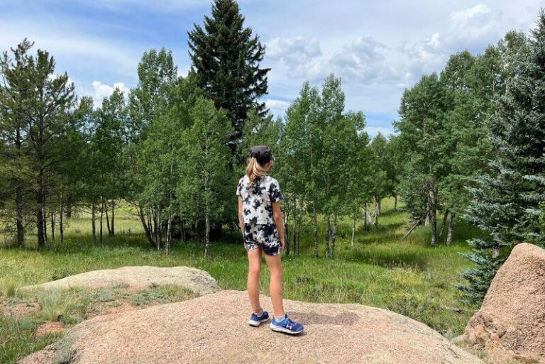 Girl in black and white tie-dye pattern shorts and shirt with black hat on. She is standing on a large boulder looking out to the grass and trees beyond