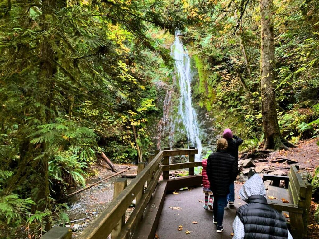 kids walking on a asphalt trail with a short wooden fence blocking the left side, a waterfall is streaming down in the distance surrounded by thick greenery trees and moss