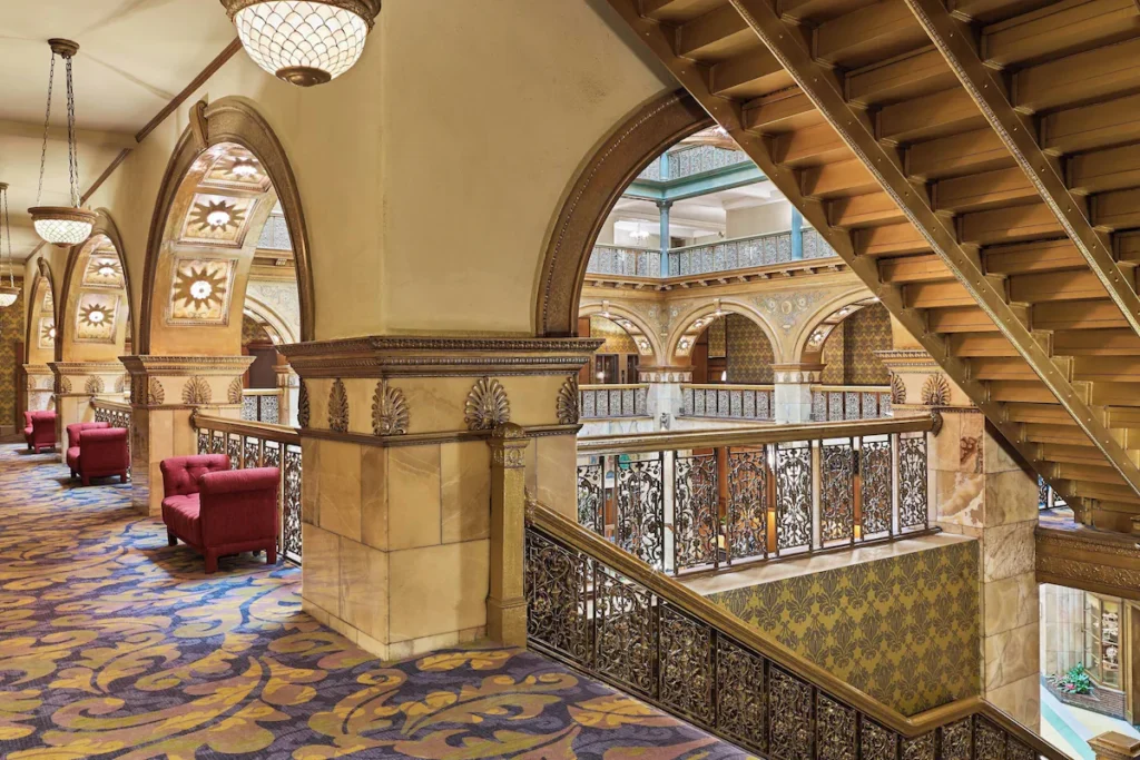 Brown Palace from an upper story in the atrium. Intricate railings through the hallway, arched ceilings, red cushioned seats along the paisley carpet
