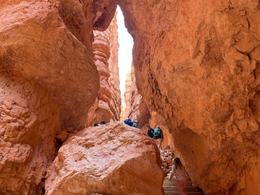 giant red rock walls close together with a giant red rock boulder in the bottom center. a Few stairs are to the right of the boulder with people going up