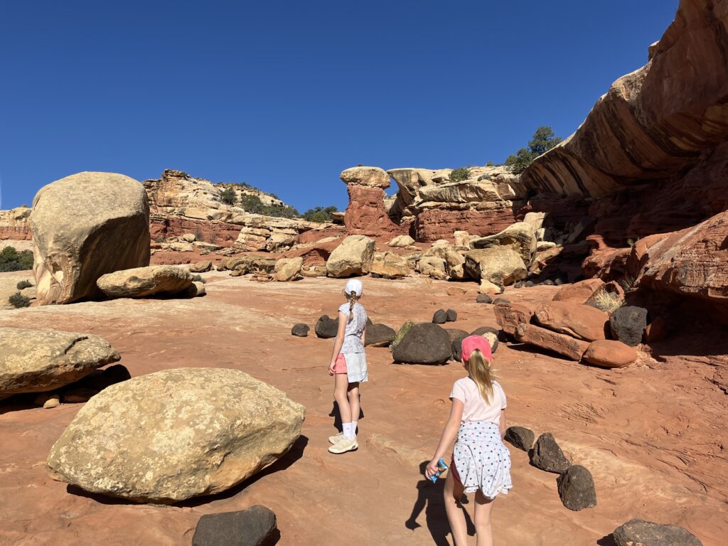 two young kids hiking across a smooth rock surface with large boulders sporadically there. Ground is reddish in color at Capitol Reef
