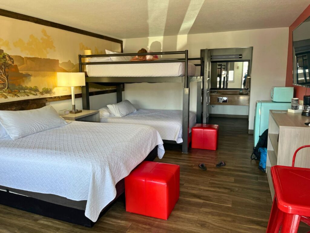 a hotel room with a double bed and a double bunk.  Red sitting blocks, a small red table nearby, and a small turquoise fridge