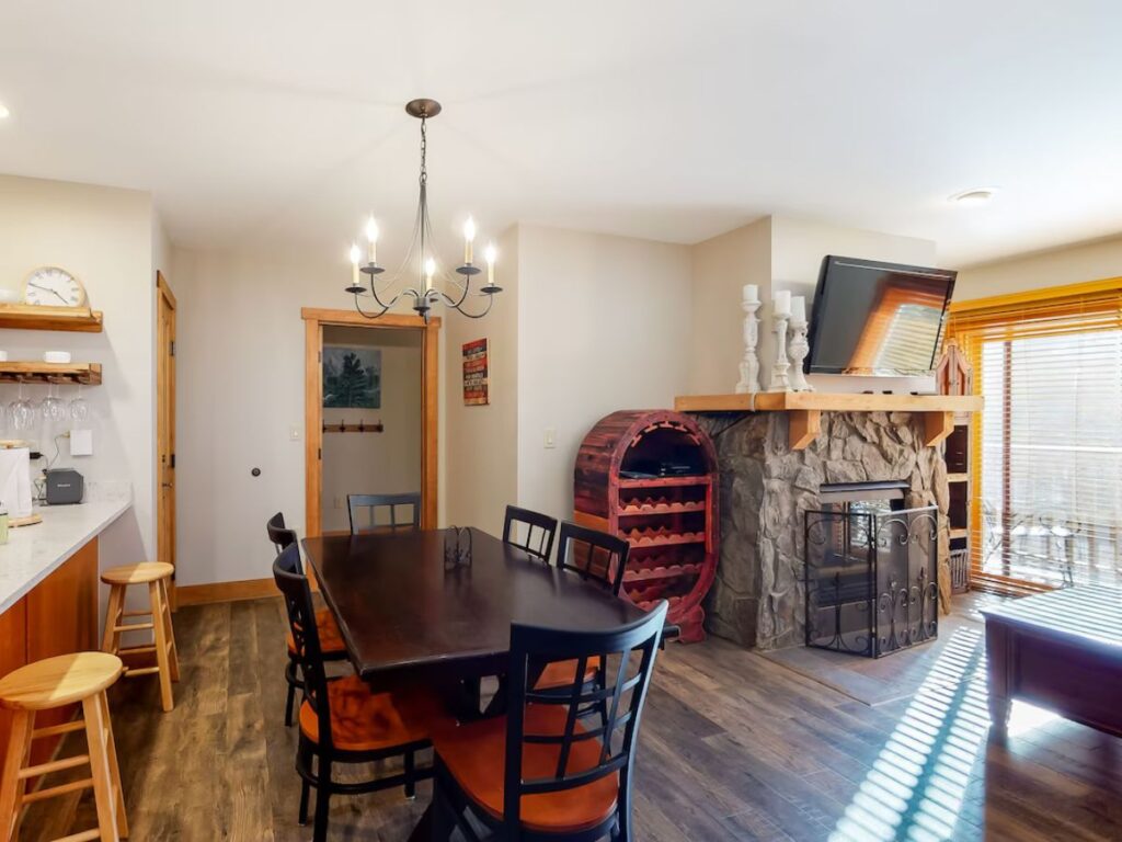 the interior of a condo in Keystone, CO. A small light fixture hangs on the ceiling above a dark brown table. to the right is a stone fireplace with a TV above. To the left is a white kitchen counter with light wooden stools