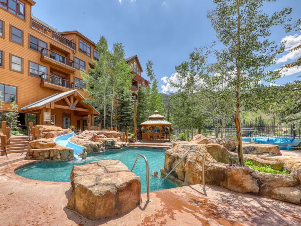a large light brown condo complex on the left with a pool area to the right. The pool has a short, gentle waterslide at ground level dropping into it. Lots of rock hardscape around the pool