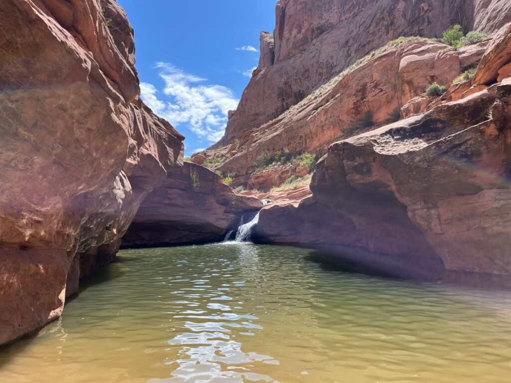mill creek falls is a great, easy hike in Moab for kids. The trail leads to this, a waterfall coming out of reddish beige rock cliffs and into greenish brown clear water below