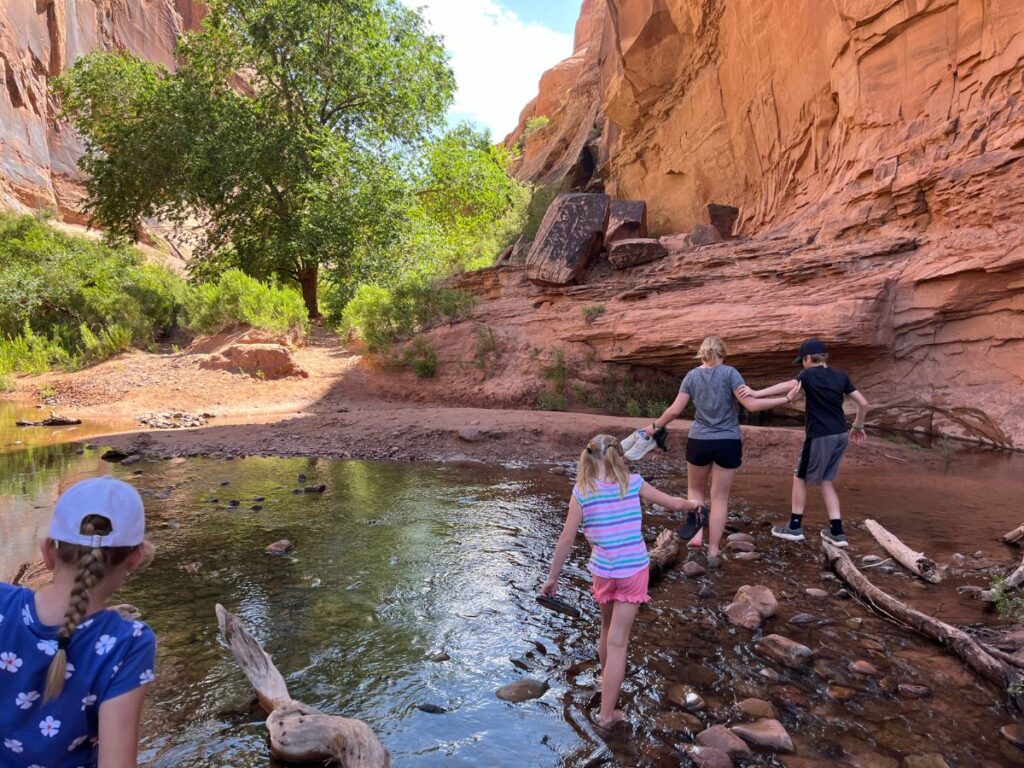 kids at a shallow river crossing. Girl in front hold the boy steady as he tries to walk on a log. Two girls behind them walk barefoot in the water. Red mountainside ahead to the right, green tree in front