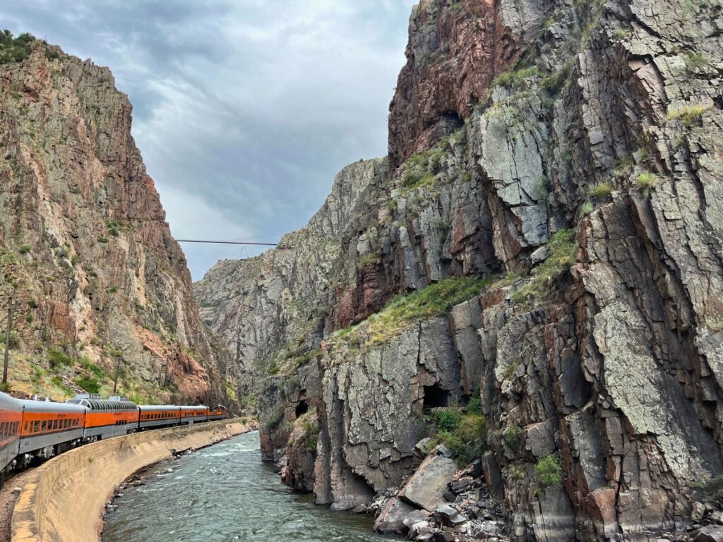 an orange train to the left running through a deep canyon along a river. A tall bridge goes across the rim ahead at the top