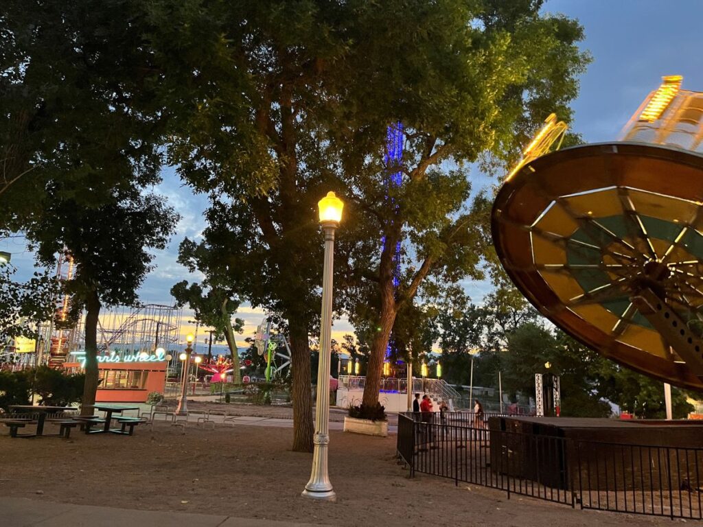 Lakeside Amusement Park at dusk. A circle ride to the right and a rollercoaster in the distance with a lamp post in the center foreground. Lakeside is on the Colorado Get Out Pass