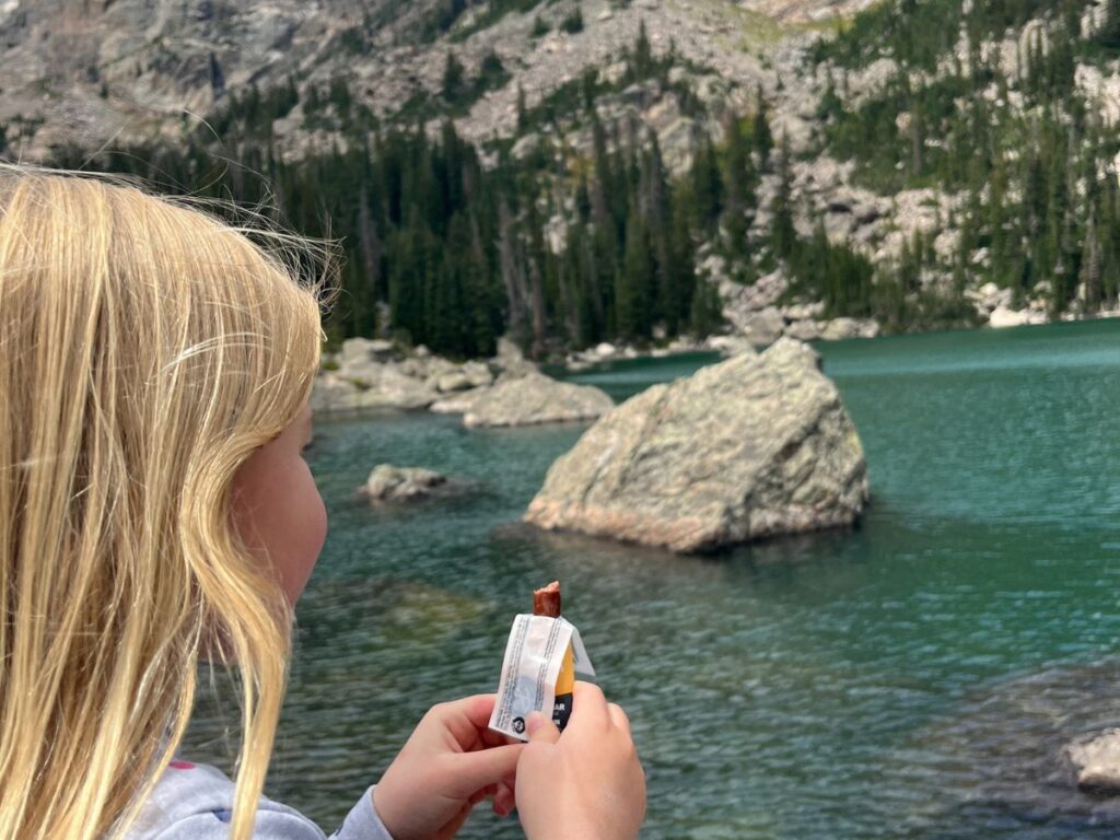 close up of a little girl 's head from the back, she is holding a small beef stick while looking out to a blue lake with a large boulder in it