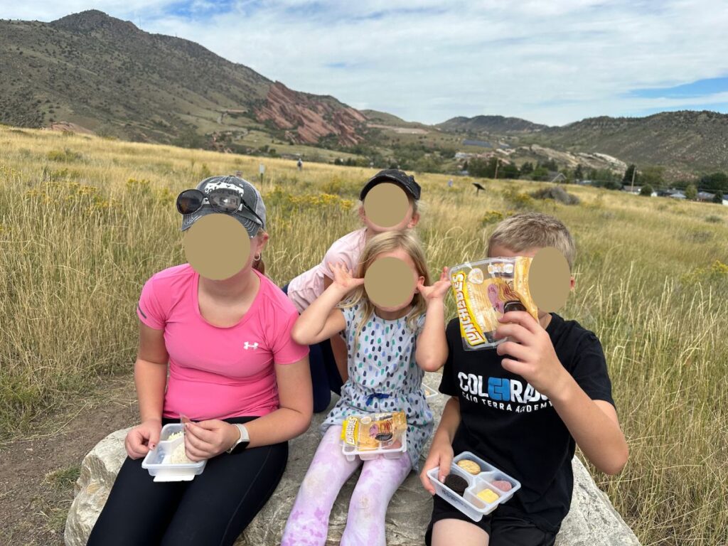 4 kids sitting with red rock formation in the distant back ground, dry grasses around them.  They are holding Lunchables as their hiking snacks