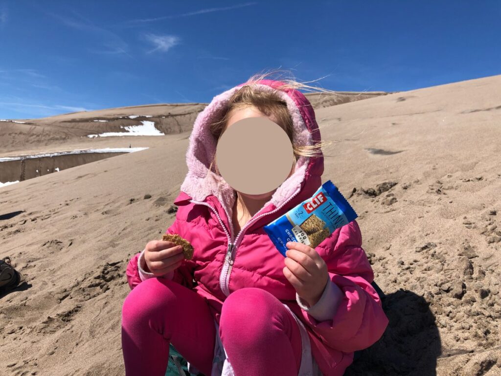 a little girl in hot pink coat and pants sits on sand holding up a cliff bar she is snacking on